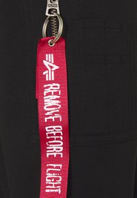 Tejido negro con un acabado texturizado, que presenta una cremallera plateada y una etiqueta de tela roja con texto blanco que dice "RETIRAR ANTES DEL VUELO".