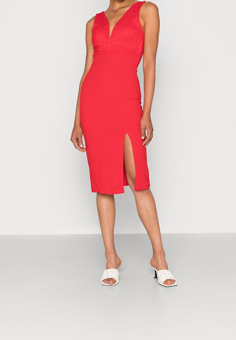 Femme portant une robe rouge sans manches avec un profond décolleté en V et une fente à la cuisse, associée à des sandales blanches à talons, debout sur un fond clair.