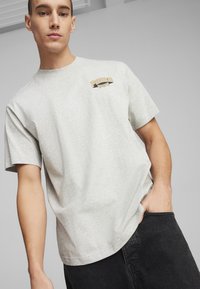 Grijze katoen T-shirt met korte mouwen, voorzien van een klein gedrukt logo in goud en zwart op het linkerborstgebied. Model draagt een donkere broek.
