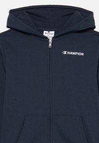 Tamnoplava dukserica sa zipom izrađena od mekog materijala, s prednjim džepom i kapuljačom. Logo "Champion" prikazan je u bijeloj boji na lijevoj strani prsnog područja.