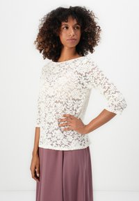 Haut en dentelle avec des manches trois-quarts, couleur blanc cassé, motif floral, texture semi-transparente, associé à une longue jupe violette.