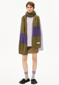 Gebreide sjaal in olijfgroen met paarse strepen, gedragen over een lavendel kabeltrui, gecombineerd met een bruine rok en meerkleurige gestreepte sokken.
