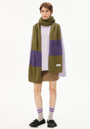 Gebreide sjaal in olijfgroen met paarse strepen, gedragen over een lavendel kabeltrui, gecombineerd met een bruine rok en meerkleurige gestreepte sokken.