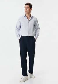 Lichtblauw gestreept overhemd met knopen, gecombineerd met navy broek. Model draagt witte sneakers, heeft de handen in de zakken en staat tegen een effen achtergrond.