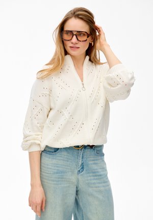 Kobieta o długich włosach poprawiająca duże okulary w oprawkach z masy perłowej, ubrana w biały dziany sweter na zamek i jasnoniebieskie, wysokowodne dżinsy.