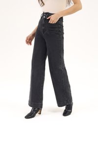 Jeans de mezclilla negros de talle alto con piernas anchas, terminados con bolsillos delanteros y un dobladillo sin rematar. Combinados con botas de tacón negro hasta el tobillo.
