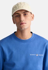 Béžová baseballová čiapka s embosovaným textom "GANT 1949". Model má na sebe modrý sveter s bielym logom "GANT 1949", okrúhly výstrih a mäkkú textúru.
