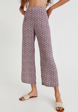 Person trägt lockere, violette Hose mit kleinem Blumenmuster und beigefarbene gewebte Sandalen, steht mit den Händen in der Hüfte.