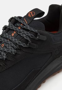 Baskets noires avec un dessus texturé, lacets gris à motifs, semelle en caoutchouc et accents orangés sur l'enseigne et les détails des lacets.
