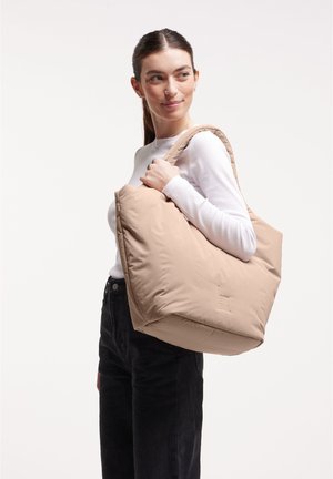 Donna con capelli scuri raccolti in una coda di cavallo, indossa una camicia bianca a maniche lunghe e pantaloni neri, portando una grande borsa imbottita beige a tracolla.