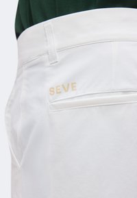 Gros plan de la poche arrière d'un pantalon blanc avec un texte beige "SEVE" et un passant de ceinture, porté avec un haut vert foncé visible à la taille.