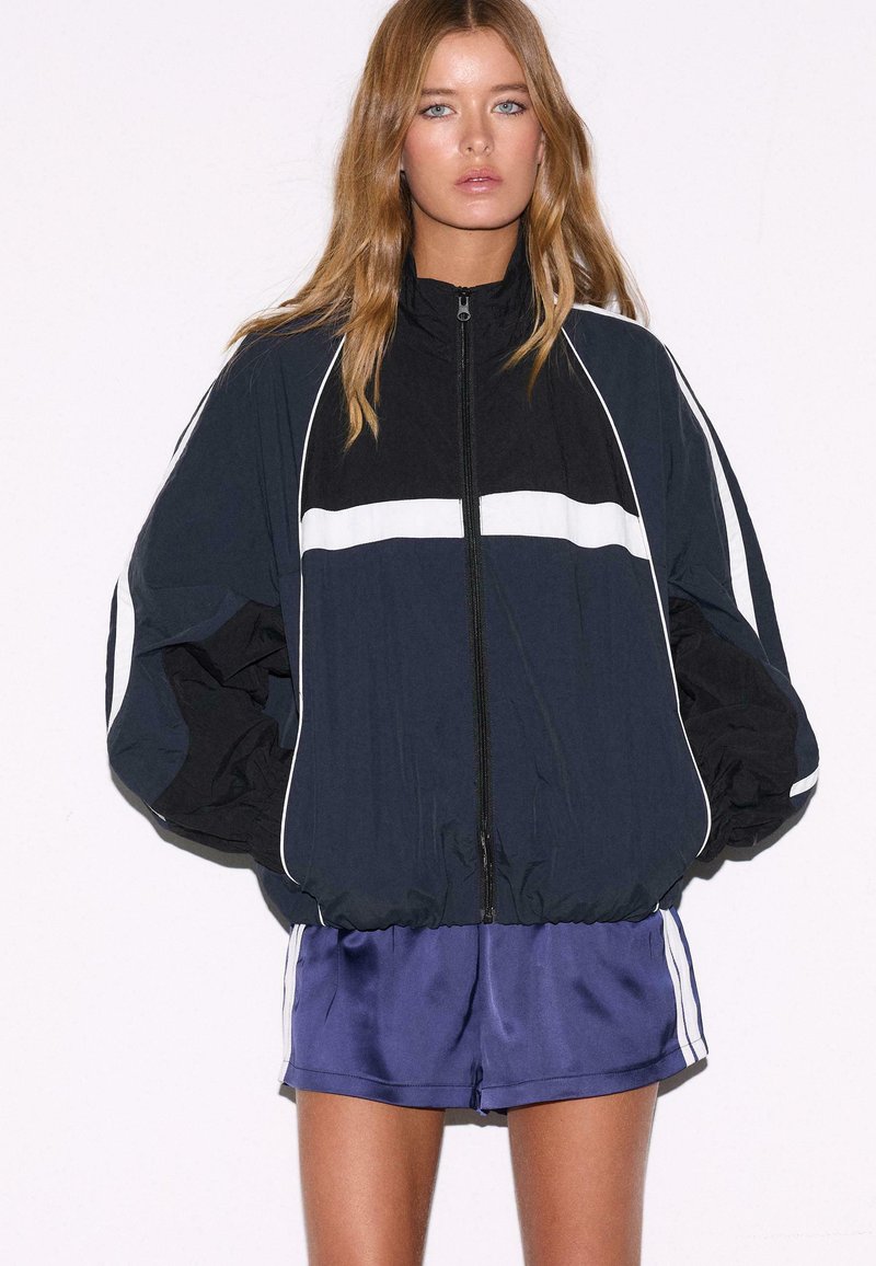 Kvinde med langt blondt hår iført en sort og navy lynlåsjakke med hvide striber og lilla træningsshorts mod en ensartet hvid baggrund.