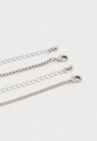 Τρία ασημένια κολιέ αλυσίδας με κλιπ αλιευτικού γάντζου και ρυθμιζόμενους δεσμούς σε σχήμα καρδιάς σε λευκό φόντο.