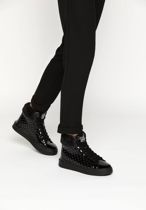 Nogi w czarnych spodniach i błyszczących czarnych trampkach za kostkę z pikowanym wzorem i futrzanym wykończeniem na białym tle.