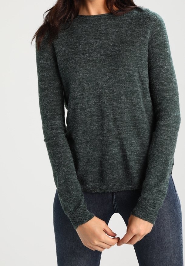 Femme portant un pull en maille à manches longues vert foncé et un jean bleu foncé, les mains légèrement jointes à la taille.