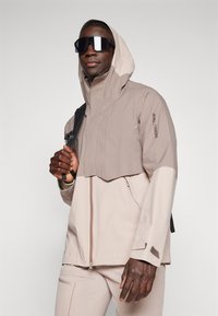 Beige waterproof jacket with a hood, featuring a cropped design and zip closure. Accented with a logo on the sleeve and a backpack strap.
