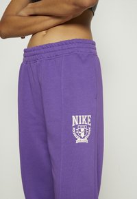 Fialové tepláky Nike s elastickým pasem, zdobené bílým logem a emblémem na levém stehně. Měkká bavlněná směs.