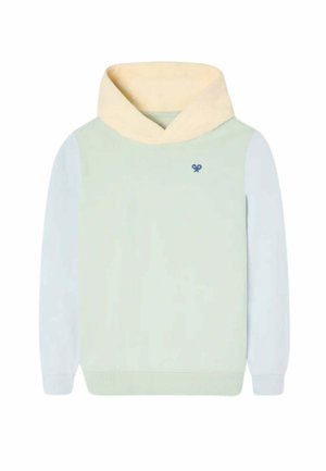 Niño de pie y sonriendo, vestido con una sudadera con capucha de color claro, pantalones cargo beige y zapatillas azules con cordones blancos sobre un fondo liso.