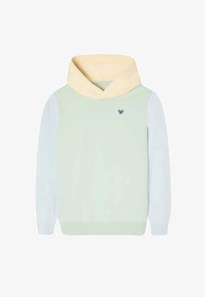 Niño de pie y sonriendo, vestido con una sudadera con capucha de color claro, pantalones cargo beige y zapatillas azules con cordones blancos sobre un fondo liso.