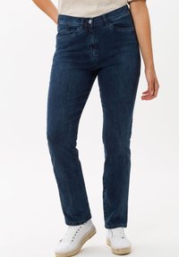 Jeans en denim bleu foncé, avec une coupe ajustée, cinq poches et une fermeture éclair. Associés à des chaussures blanches et un haut léger.