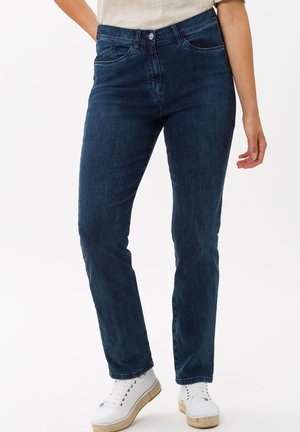 Dunkelblaue Jeans im Slim-Fit-Design, mit fünf Taschen und Reißverschluss. Kombiniert mit weißen Schuhen und einem hellen Oberteil.