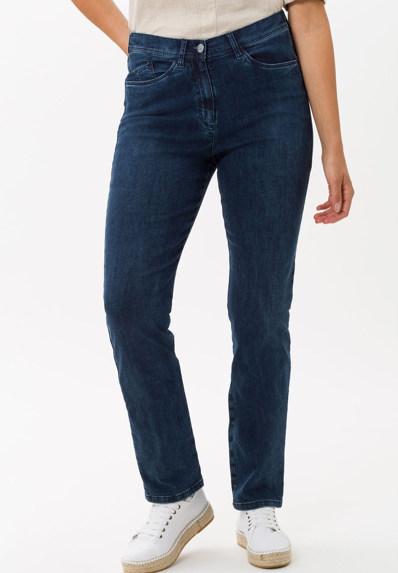 Jeans en denim bleu foncé, avec une coupe ajustée, cinq poches et une fermeture éclair. Associés à des chaussures blanches et un haut léger.