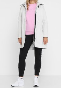 Svetle šedá waterproof bunda s kapucí, černým zipem, elastickým manžetem, v kombinaci s růžovou mikinou, černými legínami a bílými teniskami.