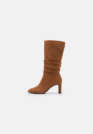 Botte marron en daim mi-mollet avec tige molle, bout pointu et talon haut et fin sur fond clair uni.