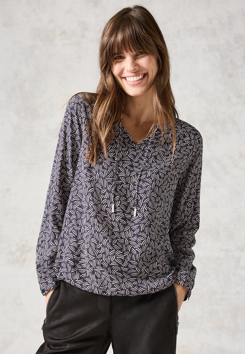 Blusa a maniche lunghe navy e bianca con motivo astratto a spirale, scollatura a V e dettagli con cordino. Indossata con pantaloni neri dal fit rilassato.