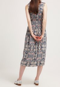 Kvinna står med händerna hopknipna bakom ryggen, iförd en ärmlös mönstrad midiklänning och silverfärgade lågklackade skor.