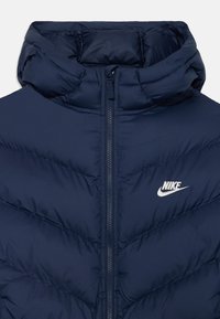 Tamsiai mėlynas pūkinis švarkas su aukštu apykakle, turintis centrinius užtrauktukus ir baltą Nike logotipą ant krūtinės. Tekstūrinis chevron raštas.