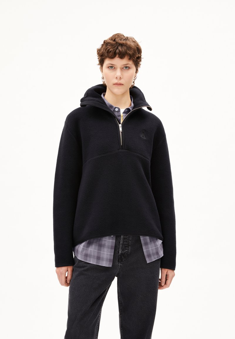 Pullover in pile nero con cappuccio, zip frontale a metà e tasca a marsupio. Indossato sopra una camicia a scacchi. Il tessuto testurizzato aggiunge calore.