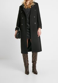 Manteau en laine noir croisé avec des boutons argentés, porté sur une robe grise. Des bottes hautes à motif serpent marron complètent la tenue.