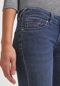 Jeans de mezclilla azul oscuro con un diseño ajustado. Las características incluyen un pequeño bolsillo delantero, costuras visibles y un acento de botón metálico.