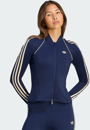 Kobieta w granatowej kurtce Adidas na zamek i dopasowanych legginsach z beżowymi paskami i logo, pozująca z rękami za plecami.