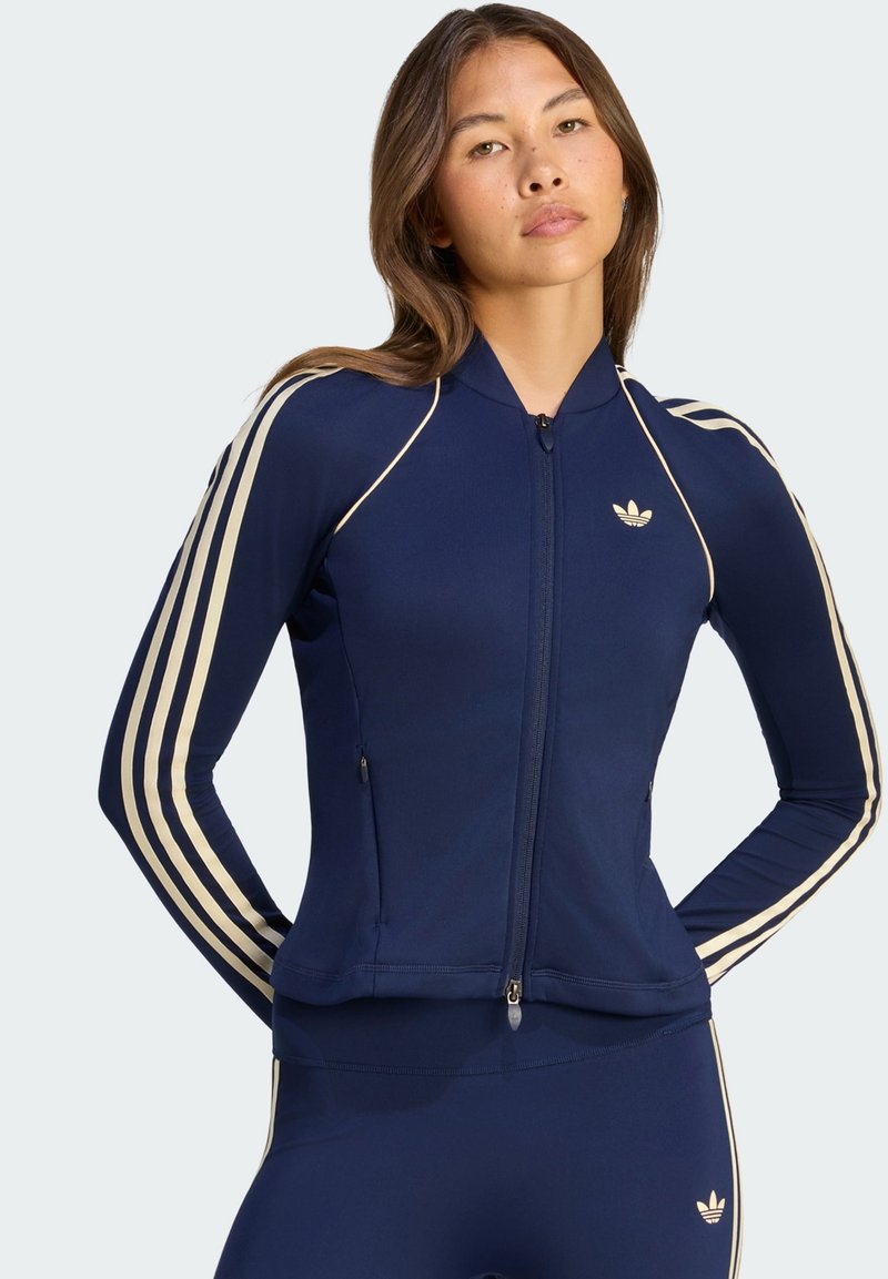 Γυναίκα με μπλε σκούρο αθλητικό μπουφάν Adidas με φερμουάρ και αντίστοιχο κολάν με μπεζ ρίγες και λογότυπο, ποζάρει με τα χέρια πίσω από την πλάτη.