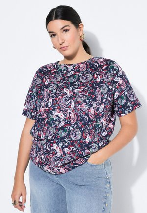 Vrouw met donker haar in een paardenstaart draagt gouden oorbellen, een marineblauw shirt met korte mouwen en paisley-patroon, en lichtblauwe spijkerbroek met gouden knopen.