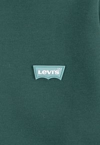 Tessuto di cotone verde con una finitura testurizzata, caratterizzato da un'etichetta del logo Levi's di colore azzurro chiaro a forma di ala di pipistrello.