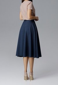 Femme portant un haut rose clair à manches courtes, une jupe midi bleu marine taille haute et des chaussures à talons beiges, debout devant un fond gris.