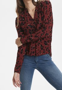 Blusa rossa a maniche lunghe con stampa leopardata nera e accenti dorati. Il tessuto ha una texture a coste e una vestibilità ampia.
