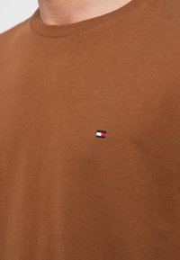 Καφέ βαμβακερό T-shirt με στρογγυλή λαιμόκοψη. Διαθέτει ένα μικρό κεντητό λογότυπο με κόκκινες, λευκές και μπλε λεπτομέρειες στην αριστερή περιοχή του στήθους.