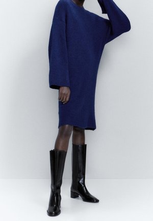 Marineblaues, oversized Strickkleid mit langen Ärmeln, getragen mit kniehohen schwarzen Lederstiefeln mit quadratischer Spitze und Blockabsatz.