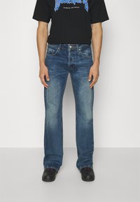 Jeans de denim azul deslavado con un diseño de pierna recta y cinco bolsillos. Llevados con una camiseta negra y zapatos negros con acentos en rosa.