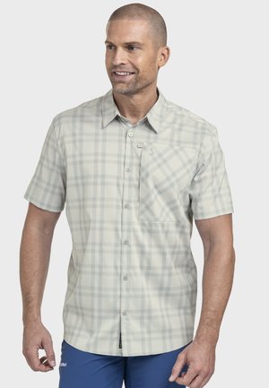 Mann trägt ein hellgraues kariertes Kurzarmhemd mit Knöpfen und Brusttasche, kombiniert mit blauer Hose, steht vor einfachem Hintergrund.