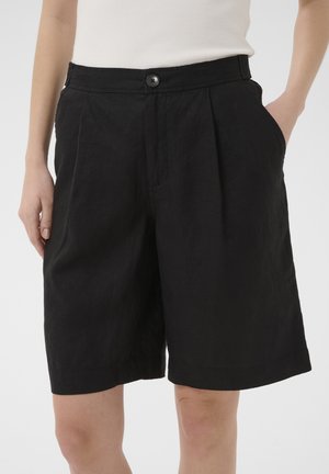 Mid-længde sorte shorts med forreste plisseringer, knaplukning og sidelommer båret af en person med hænderne delvist i lommerne.