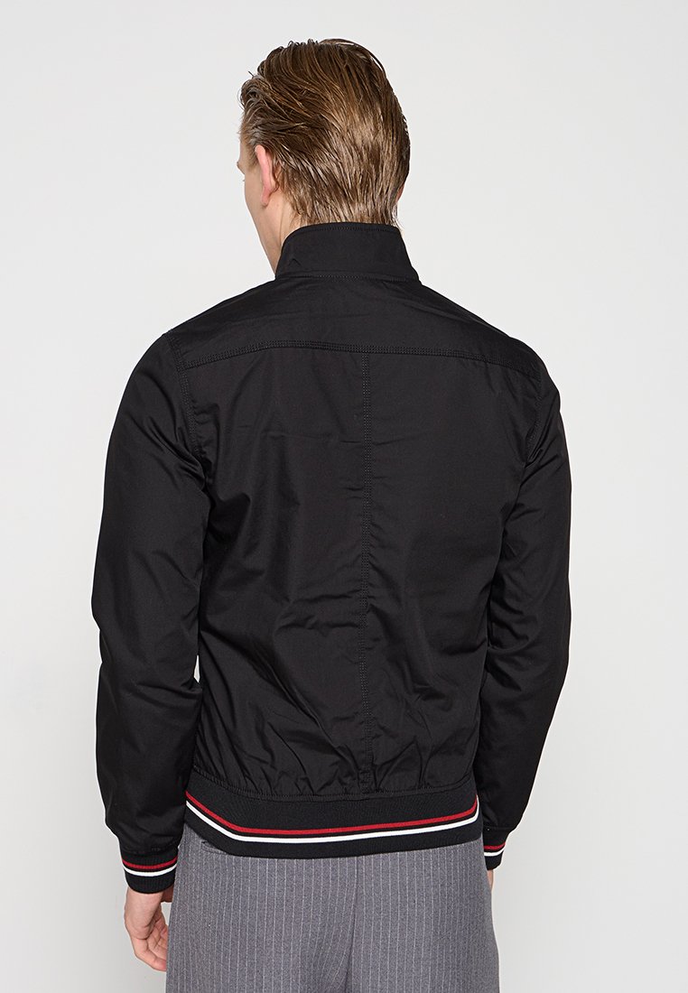 Veste bomber noire avec col montant, taille élastique avec des rayures rouges et blanches, couture diagonale dans le dos.