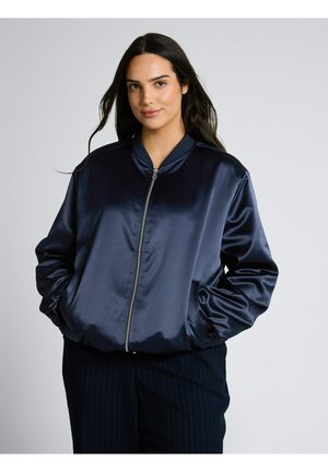 Kobieta z długimi ciemnymi włosami, ubrana w błyszczącą granatową kurtkę bomber z zamkiem błyskawicznym i rękami w kieszeniach, na jednolitym tle.