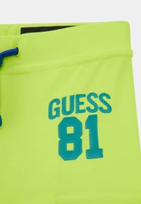 Neongröna badshorts har en blå grafisk design "GUESS 81." Tyget är glatt och inkluderar en dragsko i midjan för justering.