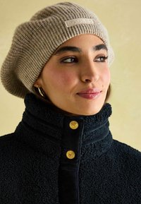 Bežová pletená bareta s prišitou etiketou; námornícky fleecový kabát s vysokým golierom a zlatými gombíkmi; hladké textúry a vzory s ryhovaním.