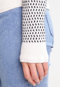 Stickad tröja med lång ärm i vitt med svart prickmönster. Ribbad manschett och synlig sömdetalj. Matchas med ljusblå denimtyg.